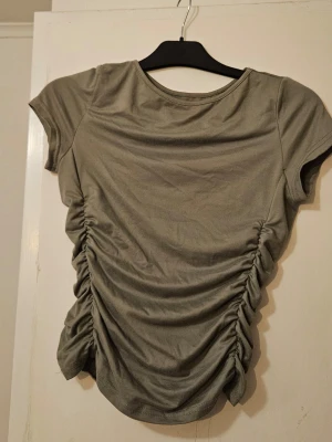 Olivgrön topp med rynkade sidor - Trendig olivgrön topp med korta ärmar och rund halsringning. Sidorna har snygga rynkade detaljer som ger en cool och modern look. Perfekt att styla med jeans eller kjol för en avslappnad vibe.