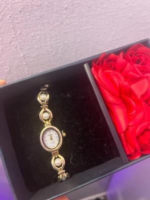 Guldig armbandsklocka med pärldetaljer - Snygg armbandsklocka från Honey i guldton med vita pärldetaljer på länken. Urtavlan är oval och vit med guldfärgade visare och index. Klockan har en elegant och feminin design som passar perfekt till dig som gillar stilrena accessoarer.