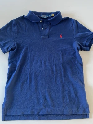 Blå pikétröja från Polo Ralph Lauren - Klassisk blå pikétröja från Polo Ralph Lauren med slim fit-passform. Tröjan har korta ärmar, krage och knappar framtill samt den ikoniska röda logotypen broderad på bröstet. Tillverkad i mjuk bomull som känns skön mot huden.