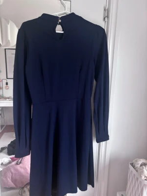 Mörkblå långärmad klänning - En stilren mörkblå klänning med lång ärm och rund halsringning. Klänningen har en lätt utställd kjol och en liten knappdetalj i nacken. Perfekt för dig som gillar enkel och klassisk stil.