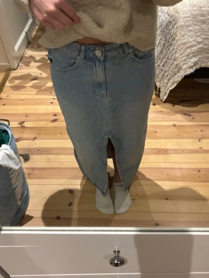 Ljusblå jeanskjol med slits - Trendig ljusblå jeanskjol från Second Female i klassisk denim med medelhög midja och en snygg slits framtill. Kjolen har femficksmodell och stängs med knapp och dragkedja. Är superskön att ha på sig men tyvärr lite för stor på mig. Aldrig använd 