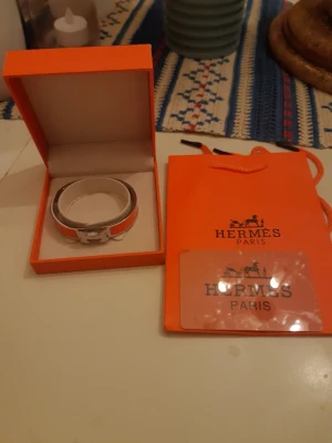 Hermès orange armband med H-spänne - Snyggt armband från Hermès i orange och silver med det ikoniska H-spännet. Armbandet har en elegant och stilren design med emaljdetaljer och levereras i originalask och påse. Perfekt accessoar för att lyfta din outfit.
