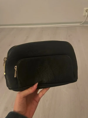 Svart necessär med gulddragkedja - Snygg svart necessär i skinnimitation med guldfärgade dragkedjor. Necessären har ett stort huvudfack och en praktisk ytterficka framtill. Perfekt storlek för att få plats med det viktigaste och enkel att ta med sig.