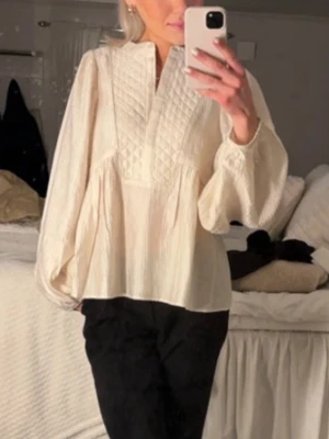 Krämvit blus från La Rouge - Krämvit blus från La Rouge med quiltad detalj framtill och knappar vid halsen. Blusen har en lös passform, rund halsringning och vida långärmade ärmar med diskreta ränder i tyget. Perfekt för en avslappnad men ändå stilren look.