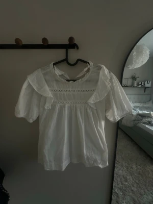 Vit blus med volang från Gina Tricot - Söt vit blus från Gina Tricot med volangdetaljer vid axlarna och puffiga korta ärmar. Blusen har dekorativ spets och rund halsringning med liten volangkant. Perfekt för en trendig och romantisk look.