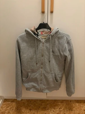 Grå zip hoodie från Burberry - Snygg grå hoodie med dragkedja från Burberry. Klassisk huva med dragsko och ikoniskt rutigt mönster på insidan av luvan och längs öppningen. Två fickor framtill och mjukt material som känns skönt mot huden. Perfekt för en avslappnad och trendig look.