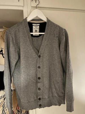 Grå Kofta  - Stilren grå kofta från Filippa K med v-ringning och knäppning framtill. Tillverkad i mjukt bomullsmaterial och har ribbade muddar vid ärmslut och nederkant. Perfekt att slänga över en t-shirt för en enkel och clean look. Har använt bara 3 gånger och tänkte sälja den. 