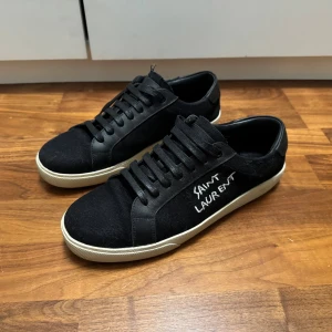 Svarta Saint Laurent sneakers - Svarta sneakers från Saint Laurent med vit broderad logga på sidan. Sulan är vit och platt, vilket ger en clean och klassisk look. Perfekta för dig som gillar stilrena sneakers. Använda en gång.