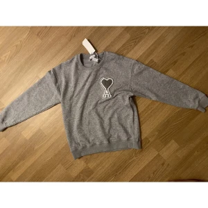 Grå sweatshirt från Ami Paris - Grå sweatshirt från Ami Paris med klassisk rund hals och lång ärm. På bröstet finns ett stort broderat hjärta med bokstaven A. Tröjan är i mjuk bomull och har ribbade muddar vid ärmslut och nederkant. Perfekt för en chill och stilren look.