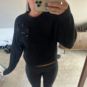 Svart stickad tröja med pärldetaljer - Svart stickad tröja från H&M i storlek XS. Tröjan har långa ärmar och är dekorerad med svarta pärlor och blommor på ena axeln. Mjuk och skön, perfekt till kyligare dagar. 