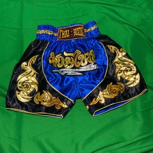 Blå Muay Thai shorts med guldmönster - Snygga Muay Thai shorts i blått och svart med detaljerade guldiga broderier och bred resår i midjan. Framtill finns en broderad text och coolt svärdmotiv. Perfekta för dig som vill sticka ut på träningen!