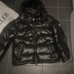 Svart Moncler pufferjacka med huva - Säljer en svart, glansig pufferjacka från Moncler med huva och dragkedja framtill. Jackan har Moncler-logga på ärmen och två snedställda fickor med dragkedja. Perfekt för kalla dagar och riktigt snygg streetstil.
