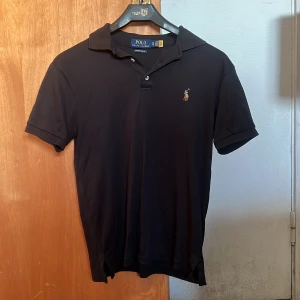  tränings pikétröja från Ralph Lauren  - Klassisk svart träningspiké från Polo Ralph Lauren i custom slim fit xs. Tröjan har korta ärmar, krage med två knappar och är perfekt för en tennis session eller en golfrunda! Hör av er vid funderingar.