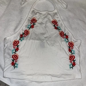 Vit broderad halterneck topp Shein - Vit halterneck topp från Shein med broderade blommor i rött, grönt och blått längs sidorna. Toppen har knyt i nacken och är ärmlös med croppad passform. Perfekt för en somrig look med snygga detaljer. Helt ny