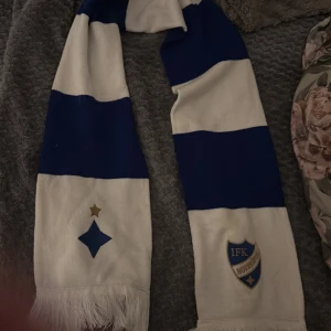 Randig IFK Norrköping halsduk - Snygg halsduk i blått och vitt med breda ränder och broderat IFK Norrköping-emblem. Halsduken är stickad och har fransar i ändarna, perfekt för att visa ditt stöd på matchen. Klassisk supporterstil med klubbens färger och detaljer.