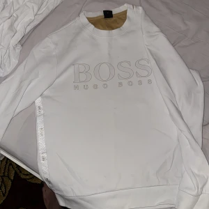Vit sweatshirt från Hugo Boss M - Vit sweatshirt från Hugo Boss med stor BOSS-logga framtill och snygga detaljer längs sidorna. Tröjan har rund halsringning, långa ärmar och ribbade muddar. Tillverkad i mjuk bomull för en clean och fräsch look.