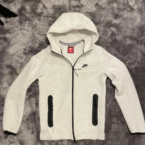 Nike ljusgrå hoodiejacka med dragkedja - Nike hoodiejacka i ljusgrått med hel dragkedja och svarta detaljer. Jackan har två svarta fickor med dragkedja framtill, huva och Nike-logga på bröstet. Perfekt för chill eller träning, riktigt clean och sportig look.