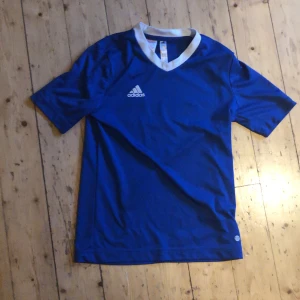Blå Adidas fotbollströja - En klassisk blå fotbollströja från Adidas med vit krage och korta ärmar. Tröjan har en enkel och sportig design med Adidas-loggan på bröstet. Tillverkad i ett lätt och ventilerande material som passar perfekt för träning eller match.