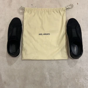 Svarta sneakers från Axel Arigato - Säljer ett par svarta sneakers från Axel Arigato med stilren design och vit platt sula. Skorna har minimalistisk look utan snörning. Perfekta för dig som gillar clean och modern stil. Medföljer original dustbag.