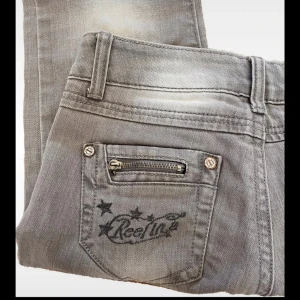 Grå bootcut jeans med dragkedja - Snygga lågmidjade grå jeans med bootcut passform. Jeansen har coola detaljer som dragkedja på bakfickan och broderad logga med stjärnor.