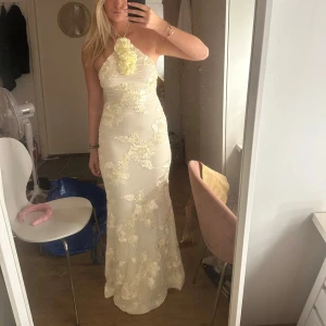 Champagnefärgad balklänning med blommor - Säljer en långklänning i champagnefärg med blommiga applikationer och halterneck med stor blomdetalj vid halsen. Klänningen är figurnära och är perfekt för speciella tillfällen som balen. Endast testats, lapp kvar