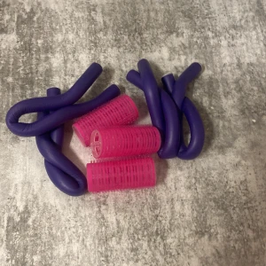 Papiljotter och andra saker  - Bilden visar hårstylingtillbehör, inklusive rosa hårspolar och lila böjbara hårspolar. Perfekt för att skapa lockar och volym utan värme. Produkterna ser ut att vara i gott skick utan synligt slitage. Passar alla hårtyper och är enkla att använda hemma.