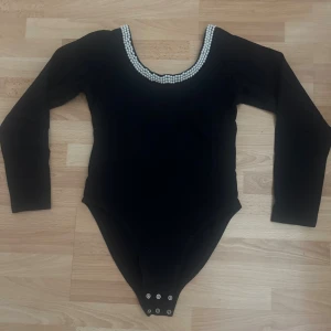 Svart långärmad body med pärldetalj - Svart långärmad body med rundad urringning dekorerad med dubbla rader vita pärlor. Bodyn har tryckknappar i grenen för enkel på- och avtagning. Perfekt för att ge en elegant touch till din outfit.