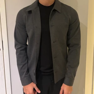 Mörkgrå overshirt med knappar - Stilren mörkgrå overshirt med klassisk krage och knäppning framtill. Modellen är 180, väger 70