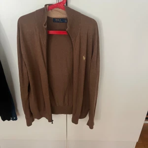 Brun kofta från Polo Ralph Lauren - Säljer en brun kofta från Polo Ralph Lauren med dragkedja framtill och klassisk logga broderad på bröstet. Tröjan har långärmad design och är tillverkad i mjukt bomullsmaterial. Perfekt för lager-på-lager och chill stil.