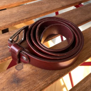 Mörkbrunt skärp i skinn med silverfärgat spänne - Snyggt och stilrent mörkbrunt bälte i äkta skinn med klassiskt silverfärgat metallspänne. Bältet är smalt och har flera hål för justerbar passform. Perfekt accessoar för att lyfta din outfit med en tidlös touch.