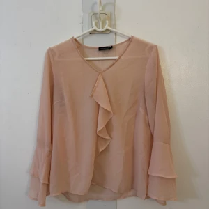 Ljusrosa blus med volang från Stockh LM - Supersöt ljusrosa blus från Stockh LM med v-ringning och volangdetalj framtill. Blusen har långa, lätt utsvängda ärmar och är gjord i ett tunt, flowy material som känns luftigt och fräscht. Perfekt för dig som gillar romantiska och feminina plagg.