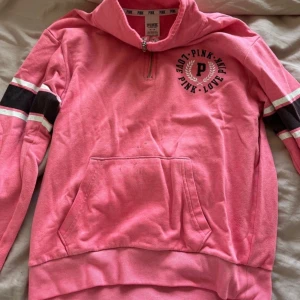 Rosa half zip hoodie från Victoria Secret  - Rosa hoodie från Victoria Secret med svart och vit logga på bröstet, svarta och vita ränder på ärmarna och praktisk känguruficka. Tröjan har half zip-dragkedja och hög krage. Perfekt för chill dagar eller när du vill ha en sportig look.