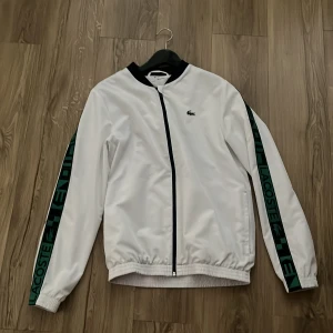 Vit vindjacka från Lacoste Sport - Snygg vit vindjacka från Lacoste Sport med svarta detaljer och dragkedja framtill. Ärmarna har gröna och svarta band med Lacoste-tryck längs hela längden. Jackan har elastiska muddar och klassisk croc-logo på bröstet. Perfekt för en sportig och clean look.