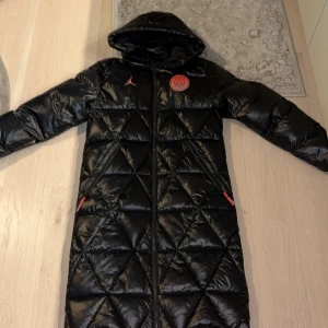 Svart lång dunjacka PSG x Jordan - Lång svart dunjacka från Jordan x PSG med glansig finish, quiltat mönster och huva. Jackan har dragkedja framtill, röda detaljer och Paris Saint-Germain-logga på bröstet samt stor PARIS-tryck på ryggen. Perfekt för kalla dagar och streetstyle. Storlek s men stor i storleken. Jag är 176 och den är i min åsikt för lång på mig. Ny pris 5000 från zalando 