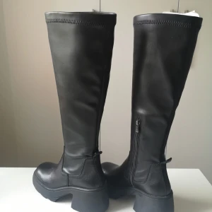 Svarta höga boots med dragkedja - Säljer ett par svarta höga boots i skinn med chunky platt sula och rund tå. Skorna har dragkedja på insidan för enkel påtagning och en stilren, minimalistisk look. Perfekta för att lyfta vilken outfit som helst.