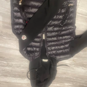 Svart hybridjacka från Moncler med huva - Svart Moncler-jacka med quiltad, glansig front och mjuka ärmar. Jackan har huva med dragsko, två dragkedjefickor och Moncler-logga på ärmen. Perfekt för dig som vill ha en stilren och sportig look med exklusiv känsla.