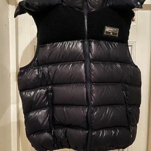 Svart dunväst från Chriztian Lane - Snygg svart dunväst från Chriztian Lane med glansig yta och detaljer i teddyfleece upptill. Västen har dragkedja framtill, två fickor med dragkedja och en stor huva med CL-logga. Perfekt för lager-på-lager under kalla dagar.