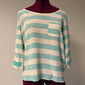 Randig silkesblus Maison Scotch - Superfin blus i 100% silke från Maison Scotch med breda mintgröna och vita ränder. Blusen har trekvartsärm, bröstficka och en snygg dragkedja bak med dekorativt hänge. 