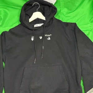 Svart Off-White hoodie med silvertryck - Svart hoodie från Off-White med vit logga och små symboler framtill. Baksidan har ett stort, unikt silverfärgat tryck i form av smältande korsade ben. Klassisk känguruficka och justerbar huva med snören. Perfekt för dig som gillar streetwear och statement-plagg.Liknande modell som originalet!