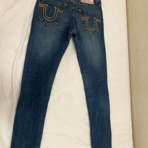 True Religion jeans, Låg midjad  - Säljer ett par blå jeans från True Religion med ikoniska gula sömmar och tydliga bakfickor. Jeansen har klassisk femficksdesign, orange kontrastsömmar och låg midja. Materialet är stretchigt denim som sitter bra längs benen.