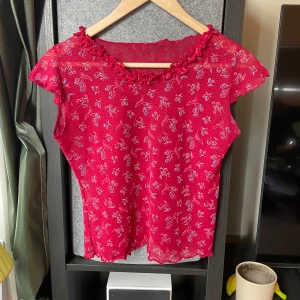 Röd blommig meshblus med volang - Säljer en röd meshblus med små blommor i blått och vitt. Blusen har korta ärmar, volangdetaljer vid halsringningen och vågig kant nertill. Tunt och lätt material, perfekt för varmare dagar.