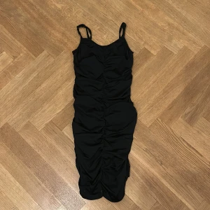Svart tight midiklänning med rynk - Svart figurnära klänning med smala axelband och snygga rynkdetaljer längs hela framsidan. Klänningen har en kroppsnära passform och är perfekt för dig som gillar en stilren och trendig look.