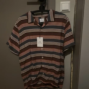 Randig skjorta från Paul Smith - Kortärmad skjorta från Paul Smith med färgglada horisontella ränder i brunt, blått, vitt och rosa. Skjortan har klassisk krage, knäppning framtill och en loose fit. Perfekt för dig som vill sticka ut med en cool och avslappnad stil.