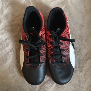 Puma fotbollsskor svart/röd mönstrad - Säljer ett par snygga fotbollsskor från Puma i svart och rött med coolt zickzack-mönster och vita detaljer. Skorna har snörning och dobbar för bra grepp på planen. Perfekta för dig som vill sticka ut på matchen!