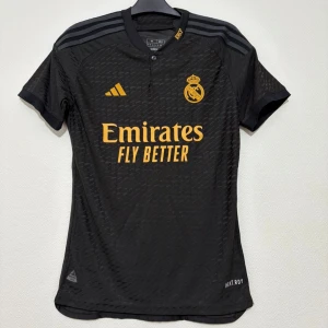 Real Madrid svart fotbollströja Adidas - Snygg svart Real Madrid fotbollströja från Adidas med gula detaljer och klubbmärke på bröstet. Tröjan har korta ärmar, rund hals och är tillverkad i ett lätt, ventilerande funktionsmaterial. Klassiska Adidas-ränder på axlarna och tryck 'Emirates Fly Better' på framsidan.