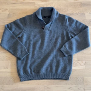 Grå stickad tröja från Zara Man - Säljer en grå stickad tröja från Zara Man med sjalkrage och en snygg knappdetalj vid halsen. Tröjan har lång ärm, ribbade muddar och är perfekt för kyliga dagar. Enkel och stilren design som funkar till många outfits.