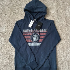 Marinblå Saint Laurent hoodie - Snygg marinblå hoodie från Saint Laurent med tryck i rött och vitt framtill. Tröjan har huva med snörning och känguruficka. Tillverkad i mjuk bomull, perfekt för en chill och stilren look.