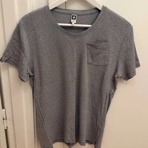 Grå t-shirt från G-Star Raw - Snygg grå t-shirt från G-Star Raw i mjuk bomull. Klassisk passform med rund hals, korta ärmar och en liten bröstficka med diskret logga. Perfekt basic-plagg med stilren look och bekväm känsla. Storlek M.