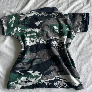 Camouflage Moncler pikéskjorta - Snygg pikéskjorta från Moncler med grön, vit och mörkblå camouflage-mönster. Klassisk krage med röd och vit rand på insidan, korta ärmar och knappar framtill. Moncler-logga broderad på bröstet. Perfekt för dig som gillar street och sportig stil.
