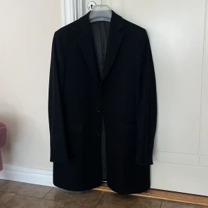 Svart kavaj från Dressmann, stl 48 - Klassisk svart kavaj från Dressmann i storlek 48. Kavajen har tre knappar framtill, två fickor med lock och en slits bak. Snyggt fodrad och med stilren passform, perfekt för dig som vill ha en enkel och tidlös look.